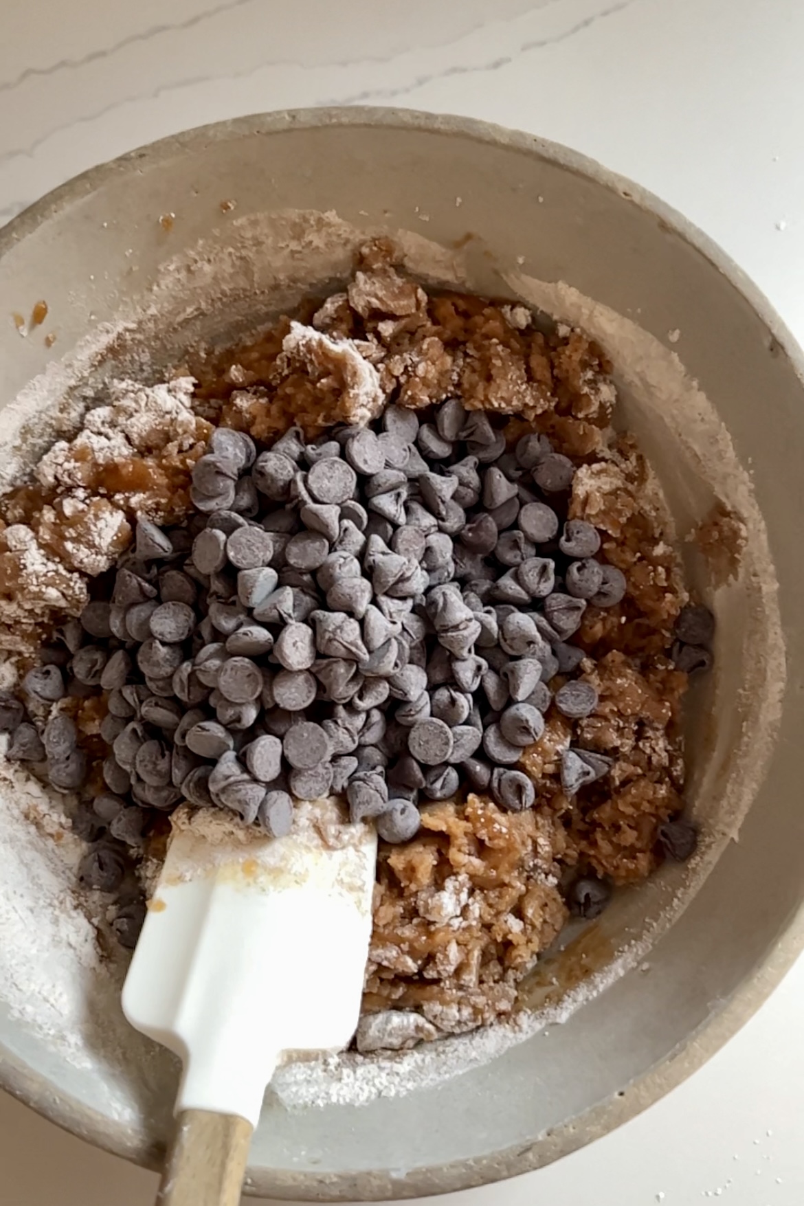 Se muestra un cuenco lleno de masa de galletas de chocolate parcialmente mezclada.
La masa contiene harina y un toque de especias de calabaza, equilibrados por una generosa cantidad de pepitas de chocolate.
Una espátula blanca con mango de madera se introduce en la masa, indicando que el proceso de mezcla está en marcha.
El cuenco está sobre una encimera clara.   