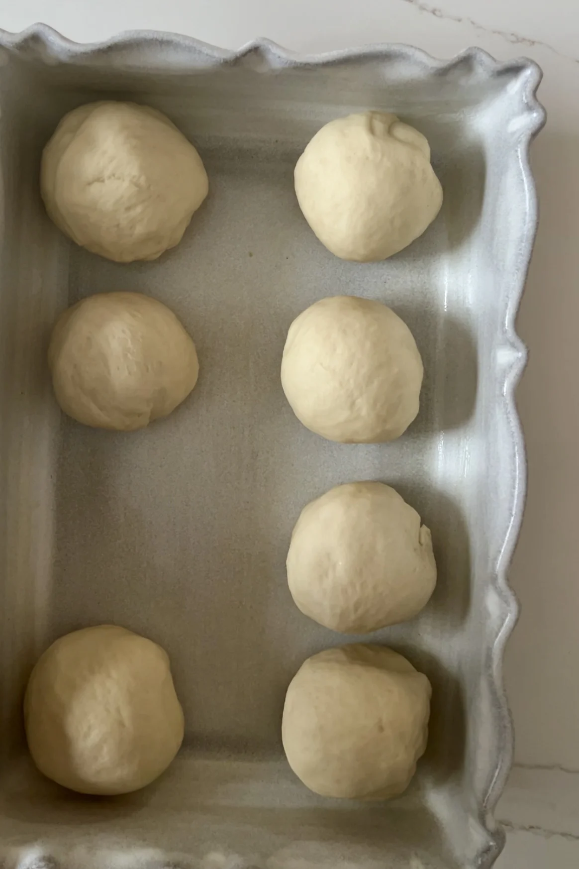 Eight dough balls are arranged neatly in two vertical columns in a rectangular, scalloped-edge baking dish. These eggless dinner rolls are evenly spaced on a light-colored surface, ready for baking into the best dinner rolls recipe you'll ever try.