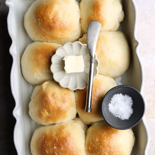 Una fuente rectangular para horno contiene seis panecillos dorados. En el centro hay un plato pequeño con una nuez de mantequilla y un cuchillo plateado para untar. Cerca hay un cuenco negro pequeño lleno de sal gruesa. El fondo es una superficie clara y texturizada.