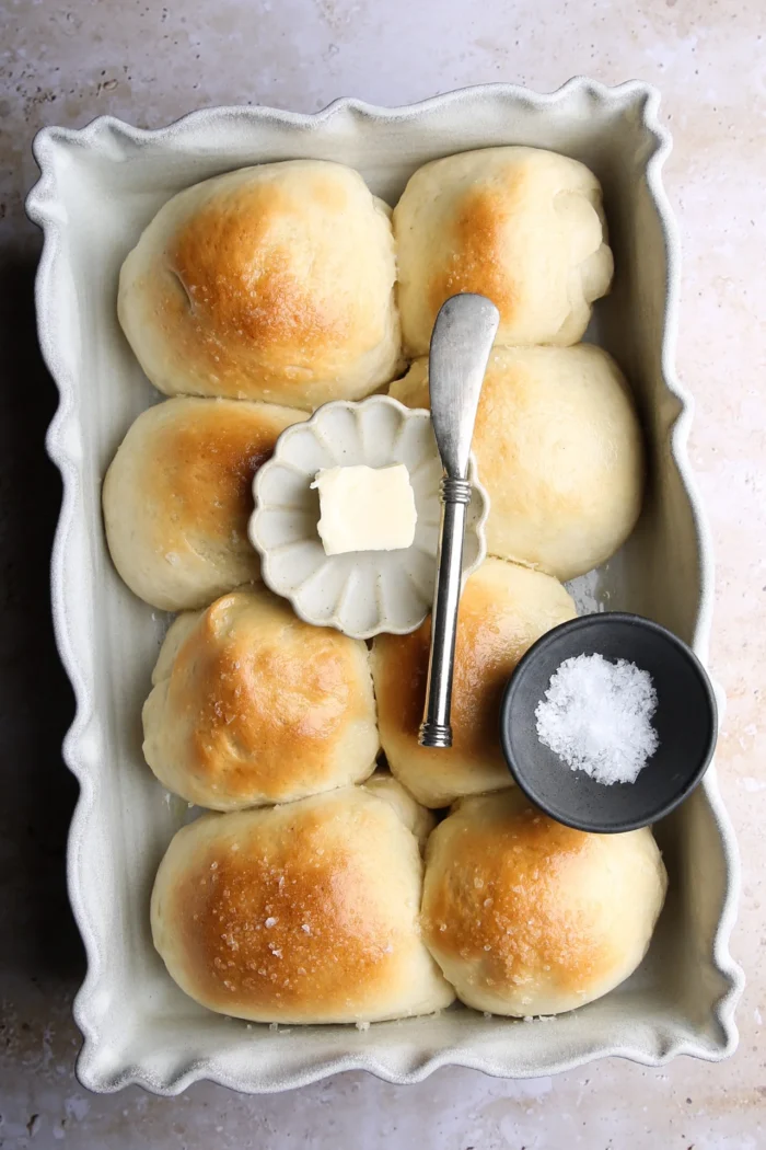 Una fuente rectangular para horno contiene seis panecillos dorados. En el centro hay un plato pequeño con una nuez de mantequilla y un cuchillo plateado para untar. Cerca hay un cuenco negro pequeño lleno de sal gruesa. El fondo es una superficie clara y texturizada.