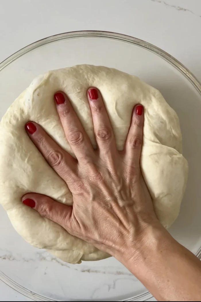 A hand with red nail polish presses down on a round ball of dough in a clear glass bowl. The dough, perfect for an eggless dinner rolls recipe, is smooth and stretches slightly under the pressure. The bowl rests on a white surface.