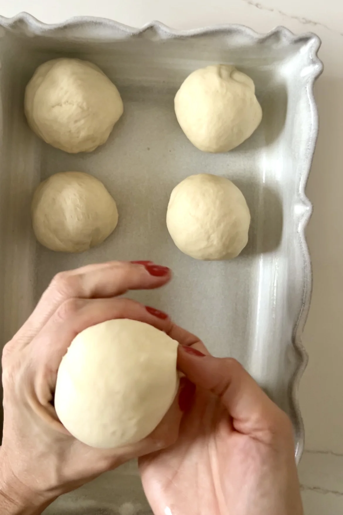 Two hands shaping a dough ball, with red-painted fingernails. Four smooth, round dough balls rest on a rectangular, wavy-edged baking dish. The scene hints at preparing eggless dinner rolls on a light-colored surface, promising a delightful dinner rolls recipe in the making.