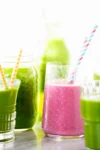 A pink smoothie in a clear glass with a blue and pink striped straw takes center stage as the ultimate smoothie. Surrounding it are various jars filled with green smoothies, each adorned with different striped straws. The bright background highlights the vibrant colors of these refreshing drinks.