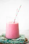 A frothy pink smoothie in a clear glass, with two colorful striped straws. The glass is placed on a stack of patterned, colorful napkins. The background is white and softly blurred, highlighting the vibrant pink of the smoothie.