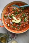 A skillet brimming with Fideo Seco, reddish-brown noodles enhanced by chipotle, is topped with avocado slices, crumbled cheese, and red onion rings. Chopped greens are sprinkled across. A blue spatula rests inside. Nearby are a drink, a small bowl of red onion, and cheese on a green napkin.