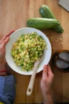 A person in a striped shirt holds a bowl of corn and chopped green vegetables, crafting a Sinaloense colache with a wooden spoon. Two zucchinis rest on the wooden table nearby, alongside a small bowl containing two gray stones.