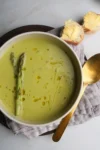 A bowl of creamy asparagus soup, made even richer with cashew milk, is garnished with whole asparagus spears and drizzled with olive oil on a wooden plate. Beside it, a gold spoon and slices of crusty bread with butter rest on a gray napkin against a smooth, light-colored surface.