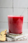 A vibrant beet juice in a clear glass on stacked terrazzo coasters. A glass straw is inserted into the juice. Slices of fresh ginger are placed beside the coasters. The background is a white tiled wall, creating a clean and minimalistic look.