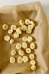 Sliced banana pieces, ready for freezing bananas, are spread unevenly on crumpled brown parchment paper. The paper rests on a light, textured surface. The circular slices reveal their soft, creamy interior with tiny seed specks at the center.