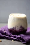 A glass filled with a layered smoothie, featuring a rich purple mixture at the bottom and a creamy, light-colored layer on top. The smoothie is placed on a crumpled purple cloth. Small nut pieces are sprinkled on top, and a gray, smooth background sets a neutral tone.