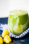 A clear glass brimming with a vibrant green smoothie, infused with matcha and swirled with creamy yellow, graces a blue surface adorned by a striped cloth. In the foreground sit three chunks of yellow mango. The softly blurred background enhances its fresh and inviting appeal.