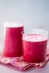 Two clear glasses filled with vibrant Pink Power Smoothies sit on a colorful floral napkin. The nutrient-packed beverages boast a frothy texture on top, with small bubbles visible. The background is softly blurred, accentuating the bright and inviting appearance of the drinks.