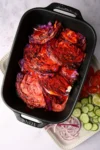 Grilled cabbage slices in a black baking dish, charred with a reddish glaze reminiscent of sarandeada sauce. Beside the dish, a white platter holds sliced cucumbers, red onions, tomatoes, and lime wedges, all arranged beautifully on a marbled surface.